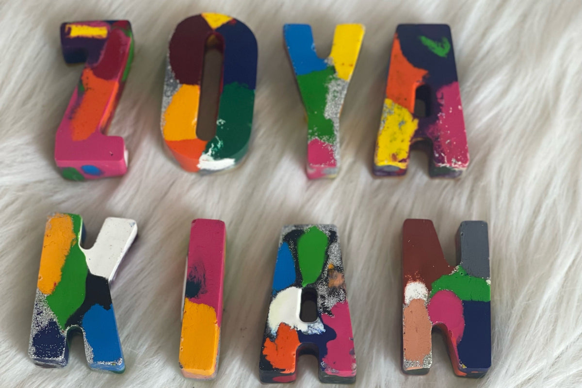 Colorful hand-painted crayon letters spelling 'JOYA KIRA' arranged in two rows on a white faux fur background.