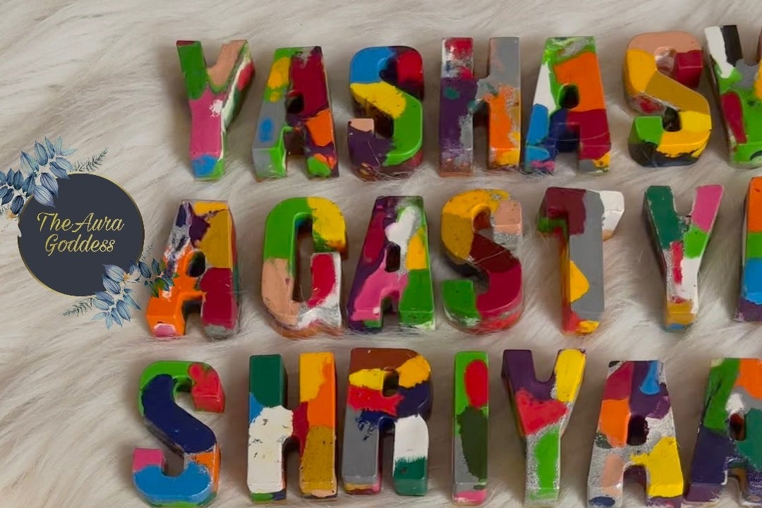 Three-dimensional crayons  letters spelling 'YASHASHW AGASTYA,SHIYAR' and 'The Aura Goddess' logo, decorated with colorful hand-made name crayons, arranged on a white faux fur background.